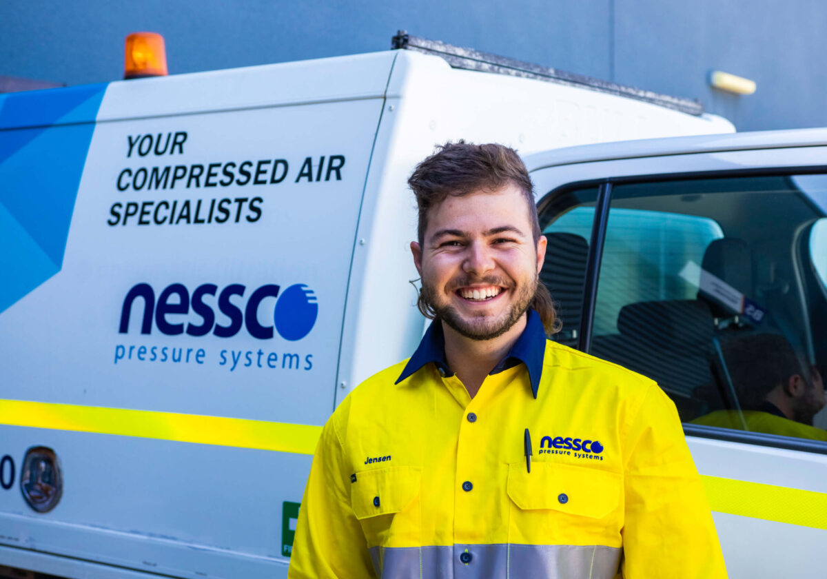 Nessco Pressure Systems (NPS) compressor service technician smiles and wears a high-vis yellow work shirt. He stands in front of a white NPS work vehicle.